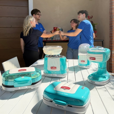 Un'inquadratura esterna su una terrazza soleggiata che presenta una collezione di piccoli elettrodomestici da cucina retrò Ariete di colore turchese brillante e bianco crema disposti su un tavolo di legno a doghe bianche. La collezione include un produttore di hot dog, un produttore di popcorn, un tostapane e un produttore di crepes, tutti con il marchio "Retro" e "Ariete" visibile. Dietro il tavolo, quattro persone (due donne e due uomini) sono in piedi, vestite casualmente, brindando con bicchieri pieni e sorridendo. Tre di loro indossano magliette blu cobalto coordinate, e uno indossa occhiali da vista. Lo sfondo mostra un edificio moderno e alcune piante decorative.