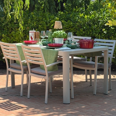 Una ripresa laterale all'aperto di un elegante set da pranzo in metallo e alluminio grigio chiaro posizionato su un patio di mattoni di terracotta. Il set include quattro sedie a doghe con cuscini sedile grigi. Sul tavolo rettangolare, è steso un runner da tavola verde texturizzato. La tavola è apparecchiata con piatti rossi e verdi lucidi, posate, calici da vino e acqua, tovaglioli di stoffa bianchi, una pentola in ceramica rossa con coperchio e, al centro, un piccolo vaso con una pianta di basilico fresco e una lampada da tavolo decorativa in metallo. Lo sfondo presenta una fitta siepe verde rigogliosa e alberi frondosi sotto la luce del sole.