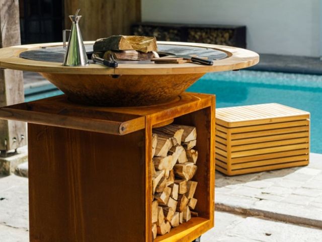 L'immagine mostra un braciere/barbecue moderno di forma circolare con una struttura in acciaio corten dall'effetto arrugginito. La parte inferiore funge da vano portalegna, ordinatamente riempito con ciocchi di legno tagliati. Sul bordo in legno chiaro del braciere sono appoggiati alcuni utensili da cucina e un'oliera in acciaio inox. Sullo sfondo si scorge una piscina dall'acqua azzurra e una panca in legno coordinata.