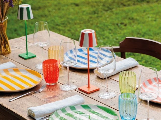 Una tavola in legno allestita all'aperto, immersa nel verde di un prato. La mise en place è caratterizzata da piatti a righe colorate (gialle, verdi, blu, rosse), calici da vino trasparenti e bicchieri d'acqua in vetro colorato. Al centro della tavola spiccano due lampade da tavolo ricaricabili senza fili con stelo sottile e paralume a righe verticali colorate (una verde e bianca, l'altra arancione e bianca).